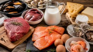 Alimentos ricos en vitamina B12 como salmón, carne de res, hígado, huevos, leche, queso, ostras y mejillones sobre una mesa de madera.